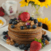 A stack of golden pancakes topped with fresh blueberries, walnuts, and a large strawberry sits on a white plate. In the background, a floral teacup and breakfast dishes are visible. A vase of bright sunflowers replaces the pineapple centerpiece, adding a summery touch. The phrase “Breakfast Made Simple – June 30” is written at the bottom.