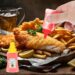 A hand squeezing ketchup from a small condiment bottle onto a piece of fried fish, served with French fries, lemon wedges, and a glass of beer in the background. Another mini bottle labeled "Tasty Honey Mustard" is placed in the foreground.