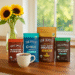 A bright, summery kitchen counter scene with a vase of sunflowers and four varieties of Four Sigmatic coffee blends arranged neatly beside a steaming cup of coffee on a rich wooden countertop.