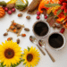 A festive fall tabletop scene featuring artificial acorns, mini gourds, pinecones, red berries, fall leaves, and two mugs of coffee, with a sunflower added for warmth and seasonal cheer.