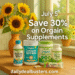 A summer-themed promotional image featuring five Orgain supplement containers on a wood kitchen counter, surrounded by fresh sunflowers and seasonal fruit. A soft golden glow filters through the sunny kitchen window, emphasizing the July 5th deal: Save 30% with Subscribe & Save at DailyDealBusters.com.