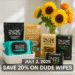 A promotional image showing a bright bathroom with a tiled countertop decorated with sunflowers in a vase. Several Dude Wipes productsβincluding Mint Chill, Shea Butter Smooth, Unscented, Odor Control, and Herbal Reliefβare arranged next to a package of Dude Underwear.