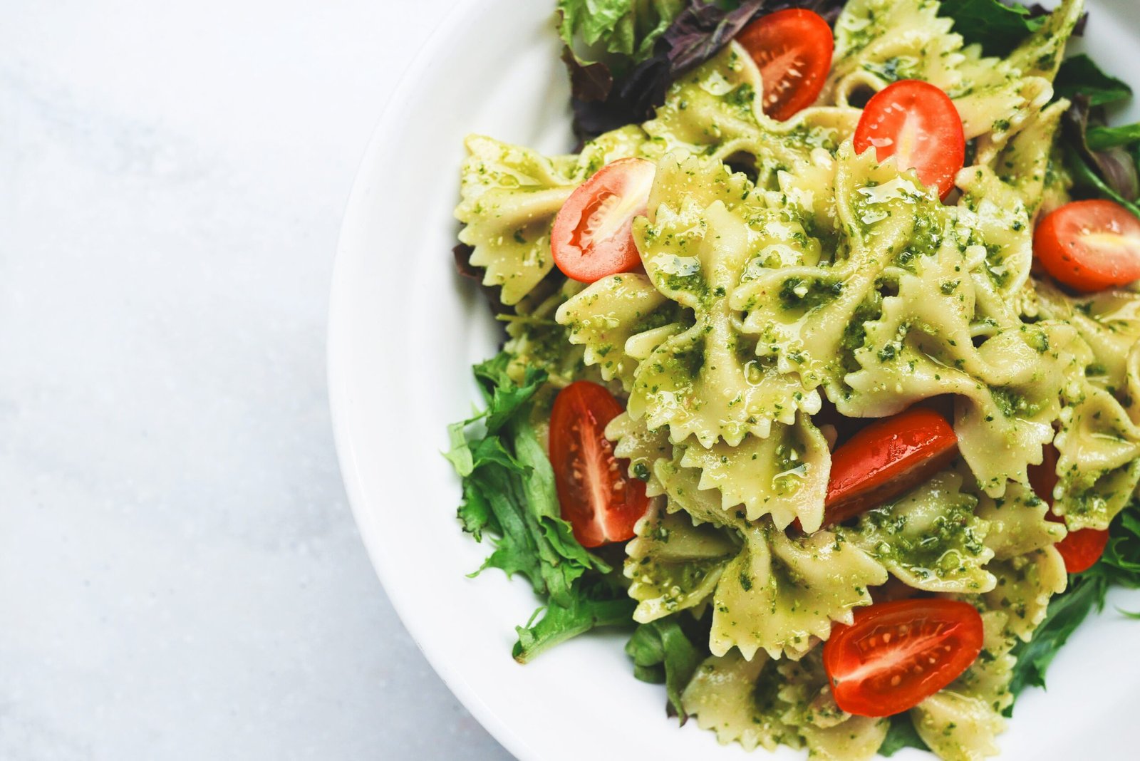 A luxurious bowl of bowtie pasta with pesto and tomatoes served on a gold-rimmed plate atop a soft white tablecloth with elegant natural lighting
